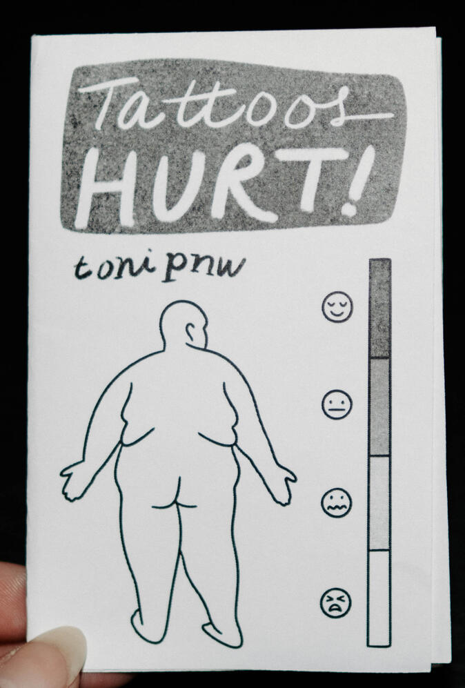 A photo of a zine cover depicting a body outline of a fat person from the back, standing next to a short, vertical pain scale with 4 faces from happy to upset. This zine is titled "Tattoos Hurt!"